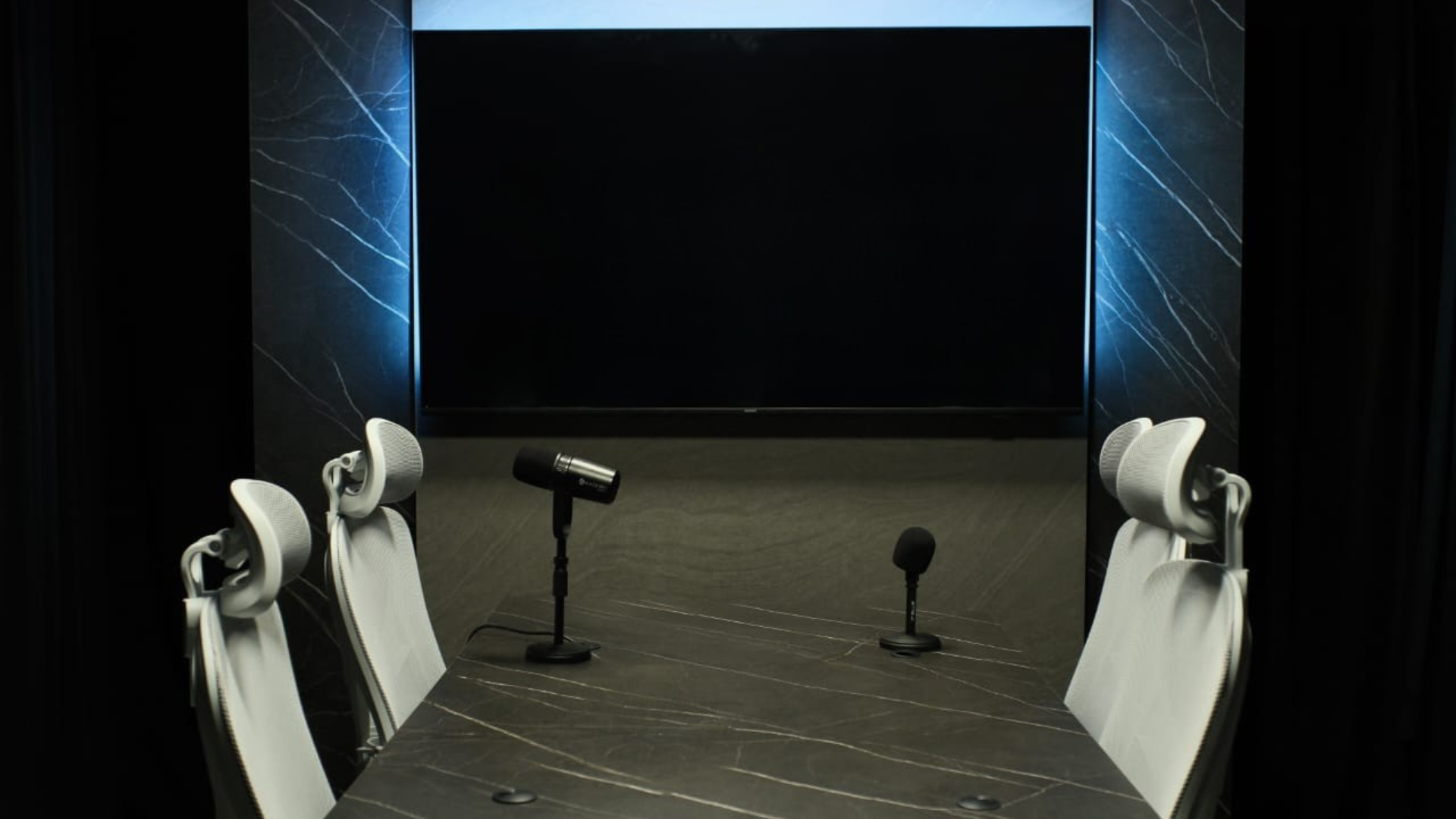 Estúdio de podcast — mesa com microfones, painel de mármore e TV
